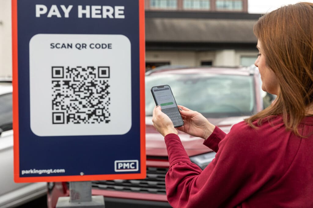 A woman scans a QR code on a "Pay Here" parking sign with her smartphone in a parking lot, with cars and a building visible in the background.
