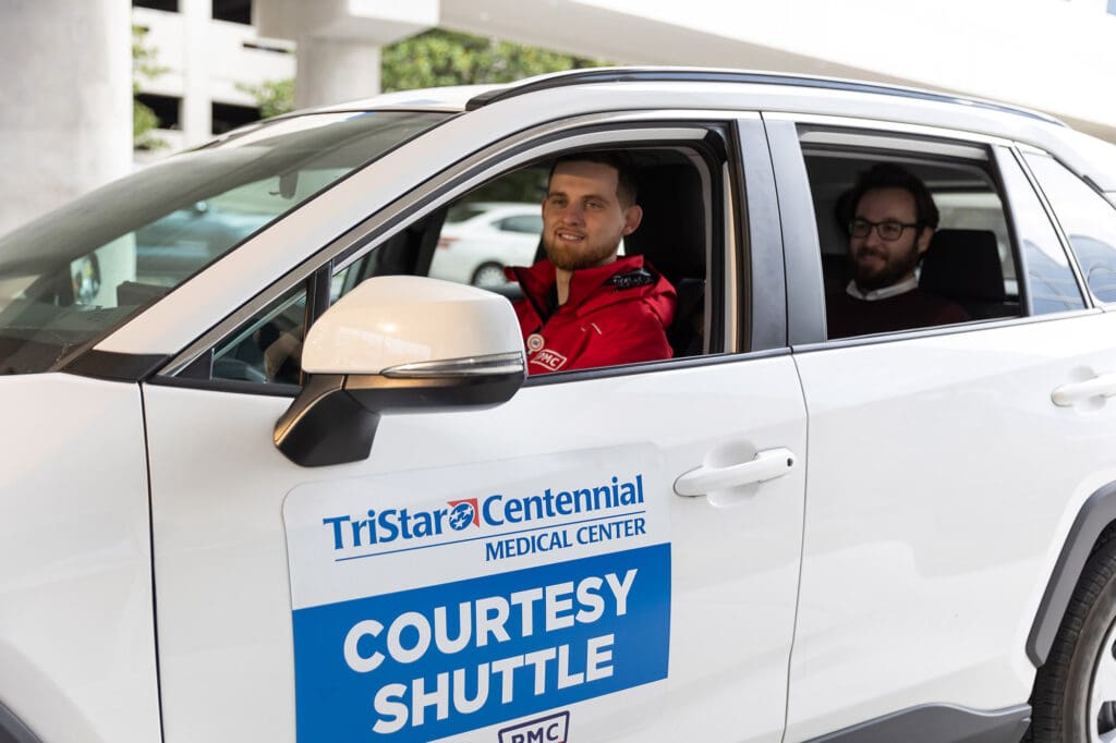 Two men sit in a white SUV labeled “TriStar Centennial Medical Center Courtesy Shuttle.” The driver is smiling at the camera, while the passenger sits in the back seat. The vehicle is parked outdoors.