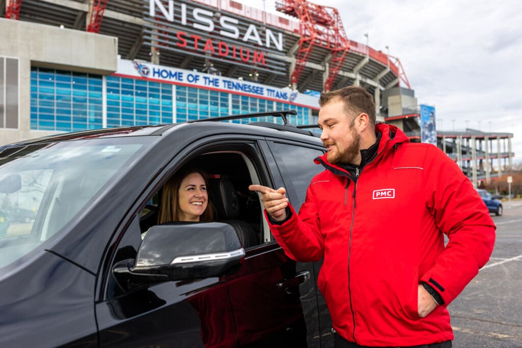 A man in a red jacket talks and gestures to a smiling woman sitting in a black SUV, with Nissan Stadium and its sign visible in the background.