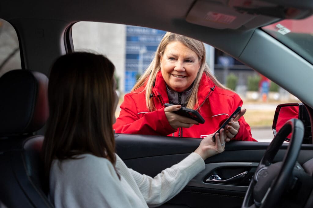 A woman in a red jacket smiles as she scans a phone held by another woman sitting in a car, seen from the passenger side window, suggesting a drive-through or curbside service interaction.
