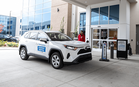 A white SUV labeled "Courtesy Shuttle" is parked outside a building entrance. A person in a red jacket stands nearby, and signs for valet parking and shuttle service are visible next to the doors.