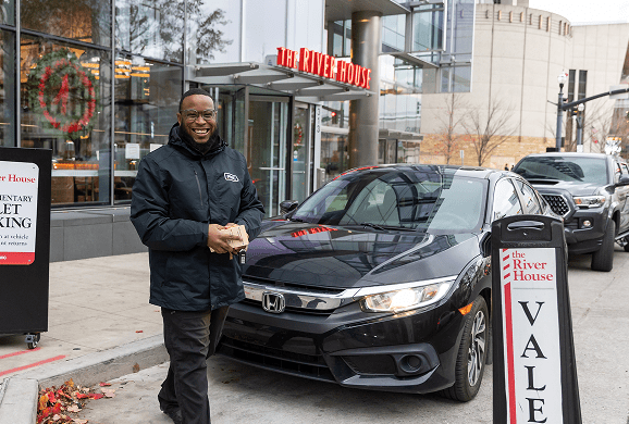 A smiling valet stands beside a parked black Honda Civic in front of The River House restaurant. A valet parking sign is visible nearby, and the restaurant is decorated for the holidays.