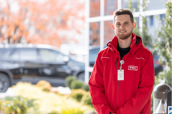 A smiling man wearing a red PMC jacket and badge stands outdoors in front of blurred greenery, a black car, and autumn-colored trees.