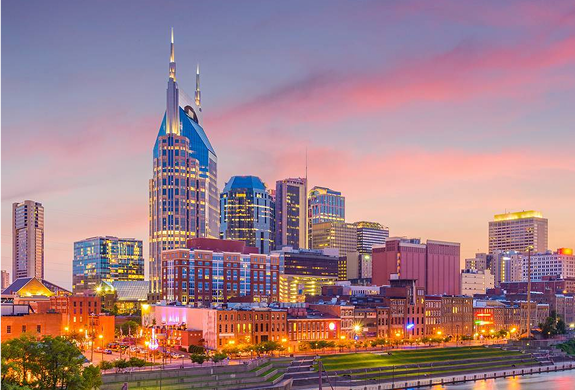Downtown Nashville skyline at sunset, featuring tall modern buildings, including the AT&T tower, with a colorful pink and purple sky and illuminated lights along the waterfront.