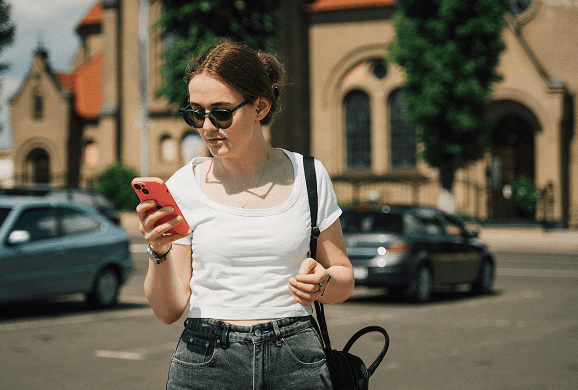 A young woman wearing sunglasses and a white t-shirt looks at her phone while walking through a sunny parking lot with parked cars and buildings in the background.