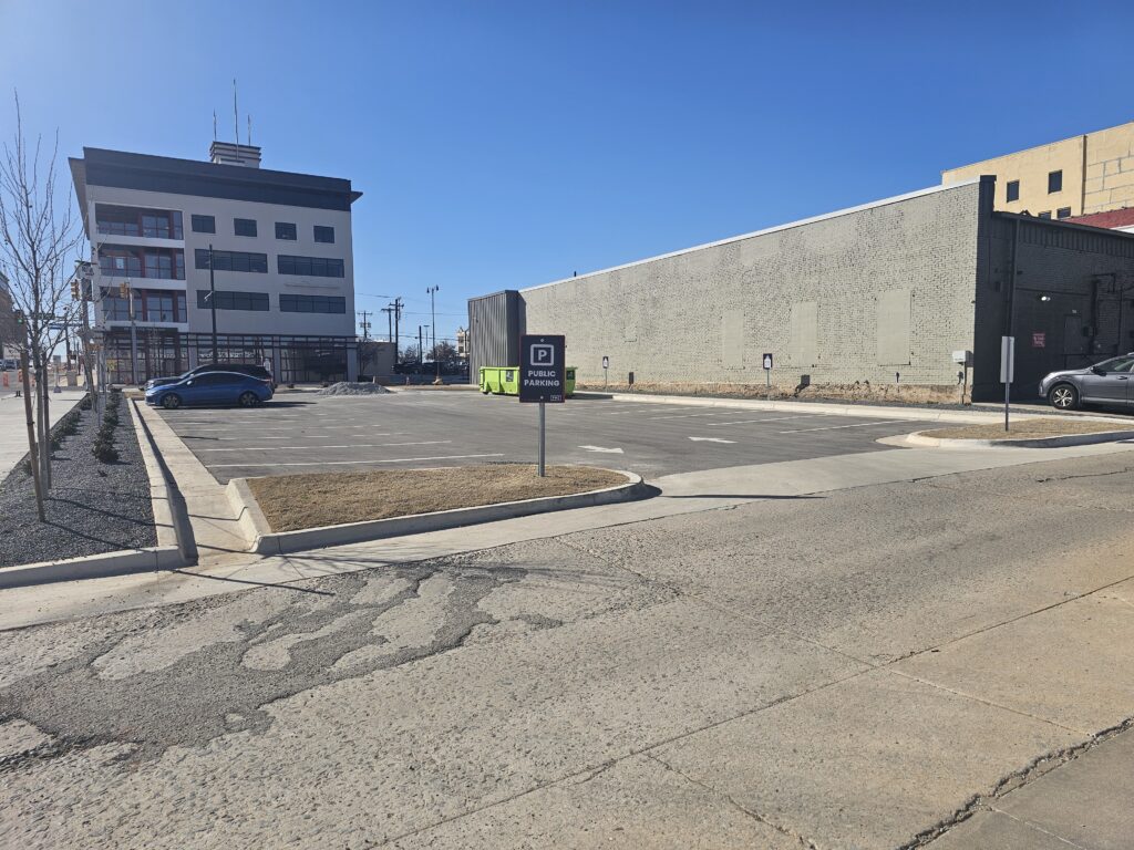 A nearly empty parking lot with a few parked cars, a sign for public parking, and a modern building with large windows in the background under a clear blue sky.