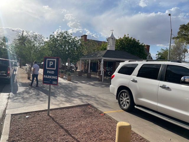 A parking lot with a "Paid Parking" sign, several cars parked, a person walking, and a small building with a cupola and trees in the background on a sunny day.