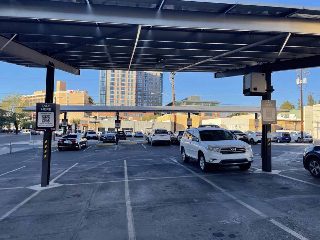 A parking lot with several cars parked under a canopy, PMC signs with QR codes for payment visible, and city buildings in the background.