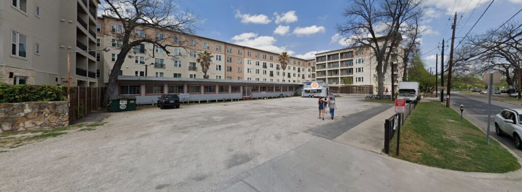 A wide gravel parking lot with a food truck, a few parked cars, and two people walking. Large apartment buildings and leafless trees line the background under a partly cloudy sky.