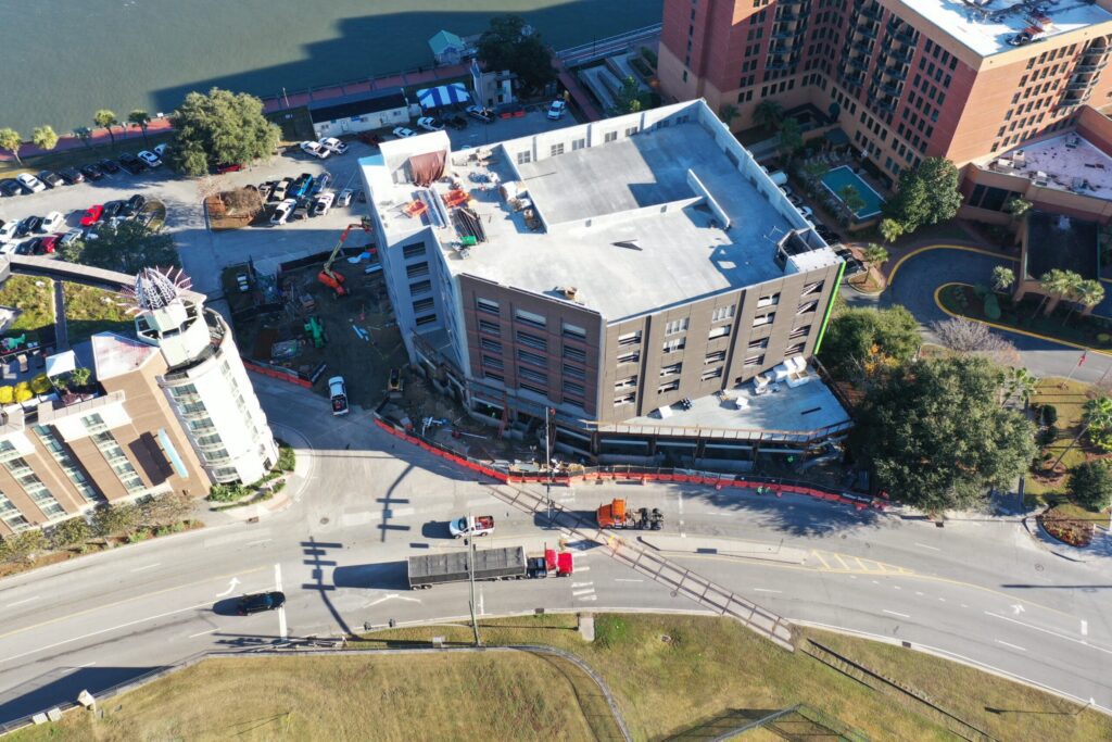 Aerial view of an urban area featuring a multi-story building near a curved road. Construction vehicles and equipment are present, and other buildings, trees, and a body of water are nearby.