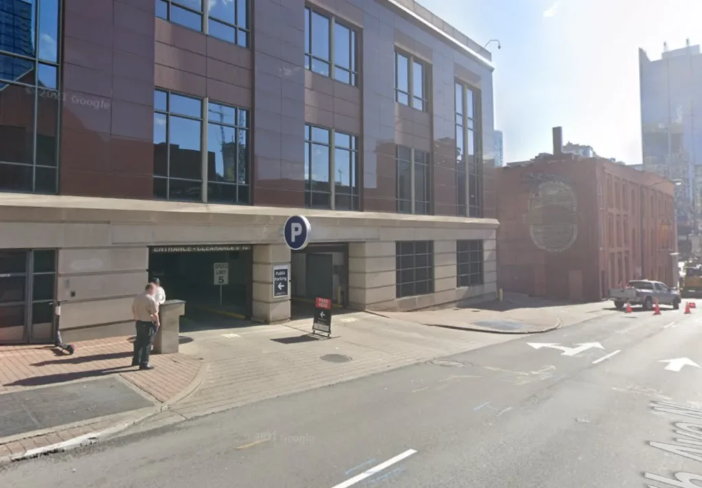 A man stands at the entrance of a parking garage in a modern building on a sloped city street. A parking sign is above the entrance, and traffic cones are set up further down the street.