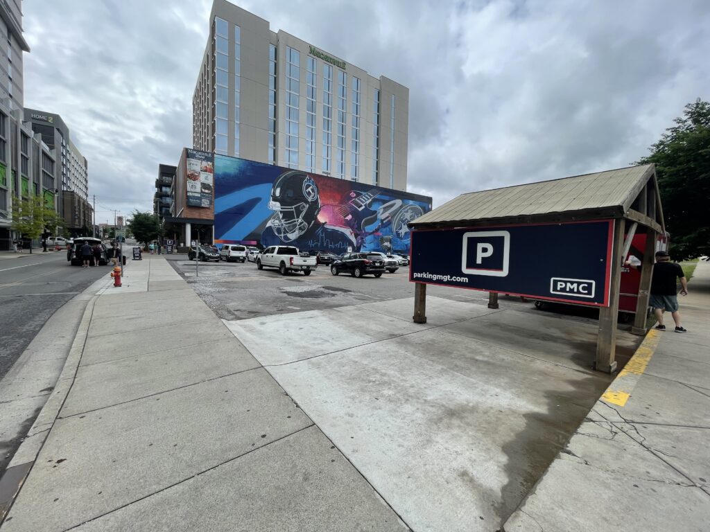 A city street with a parking lot in the foreground, a large mural featuring sports figures on a building wall, several parked cars, and tall buildings under a cloudy sky.