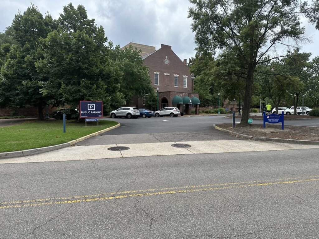 A brick building with green awnings and arched windows is surrounded by trees and parked cars. A sign on the lawn reads "Public Parking." The sky is cloudy and the street is in the foreground.