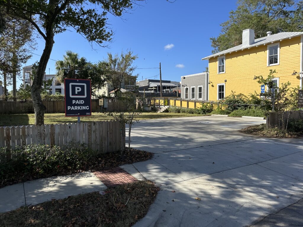 Parking Management Company parking lot with a "Paid Parking" sign and PMC logo, surrounded by a wooden fence and greenery. A bright yellow building is on the right, under a clear blue sky with a few scattered trees in the background.