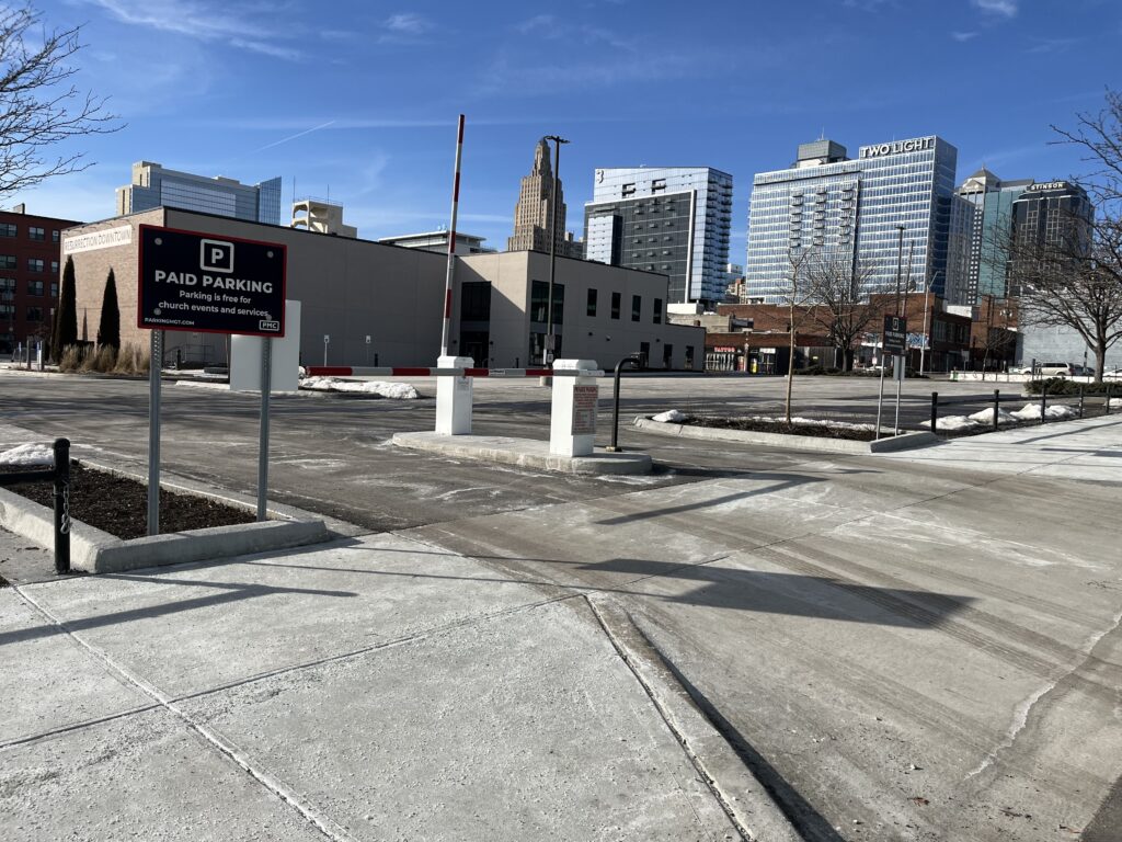 A nearly empty paid parking lot with a sign in the foreground, surrounded by buildings and city skyscrapers under a clear blue sky. Some patches of snow remain on the ground.