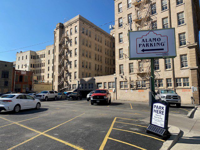 A sunny urban parking lot with multiple cars parked. A large sign reads "Alamo Parking" and a smaller sign below says "Park Here." Tall beige buildings with fire escapes stand in the background.