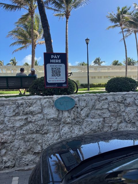 A parking pay sign with a QR code stands on a sidewalk next to a stone wall. Two people sit on a bench under palm trees, and a bicycle is nearby. A parked car's hood is visible at the bottom of the image.