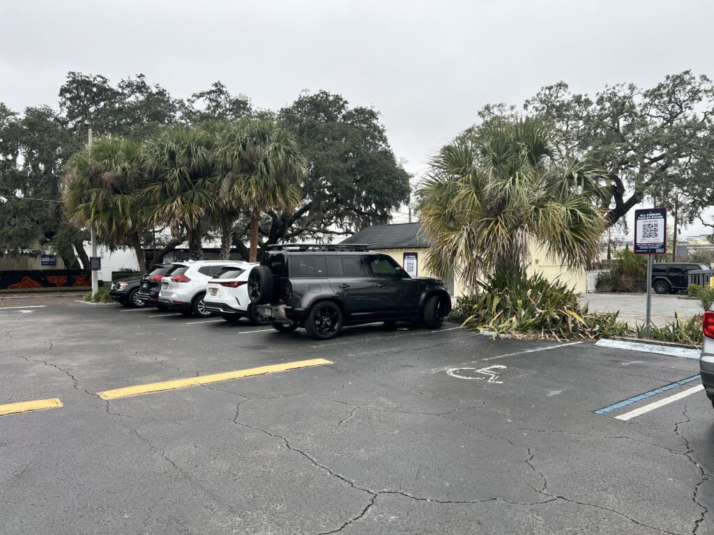 A parking lot with several cars parked, including a black SUV next to palm trees. There is a building in the background and a visible handicapped parking space in the foreground. The sky is overcast.