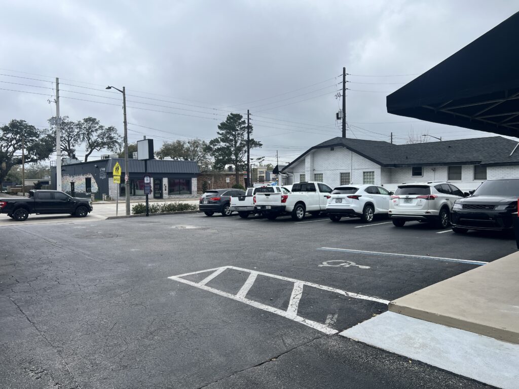 A parking lot with several parked cars on a cloudy day. In the background, there are some buildings, trees, and utility poles. A handicapped parking space is visible in the foreground.