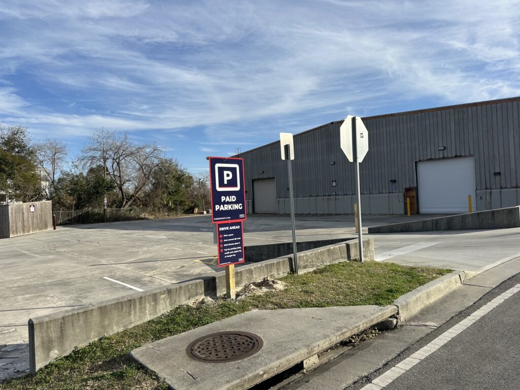 Parking Management Company parking lot with a PMC sign in front, next to a large gray warehouse building. A stop sign and another street sign are also visible.