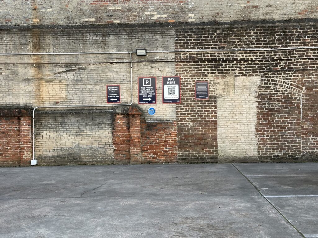 Parking Management Company-owned parking lot in front of an old brick wall with several PMC parking signs, including a "Pay Here" sign with a QR code, mounted on the wall.