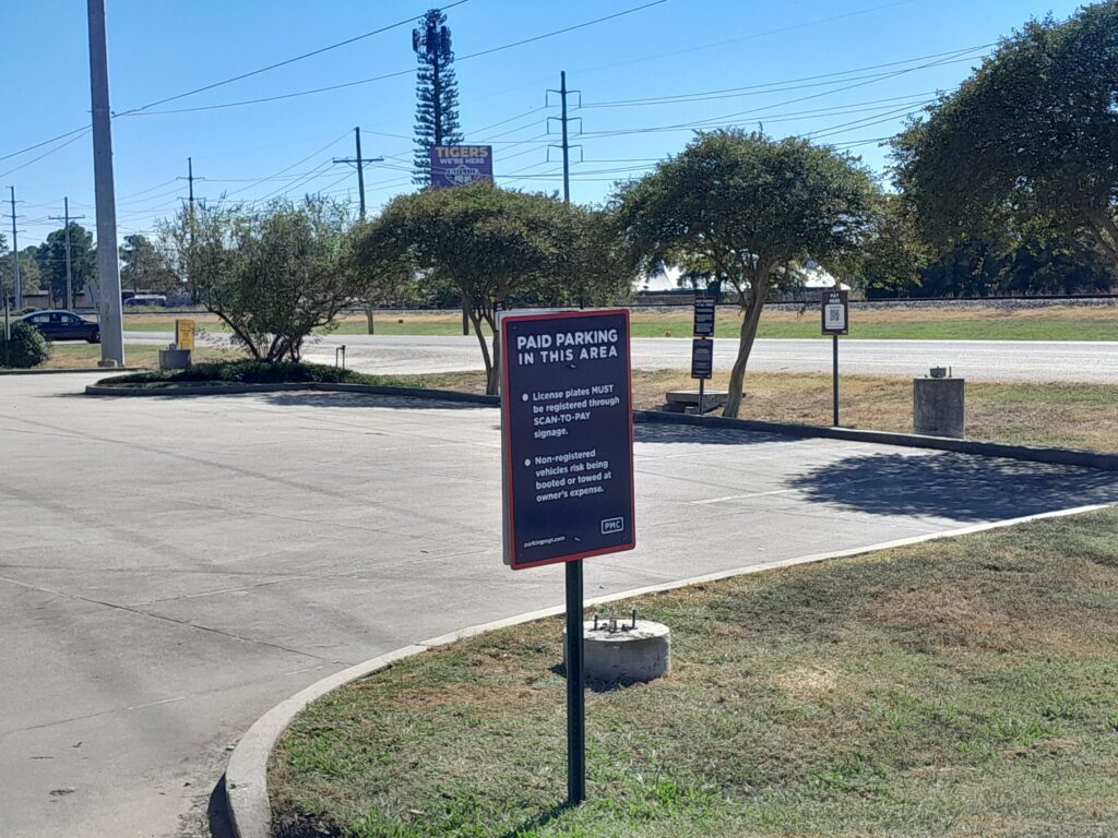 A sign in a nearly empty parking lot reads “Paid Parking in This Area.” Trees, a road, and a billboard are visible in the background under a clear blue sky.