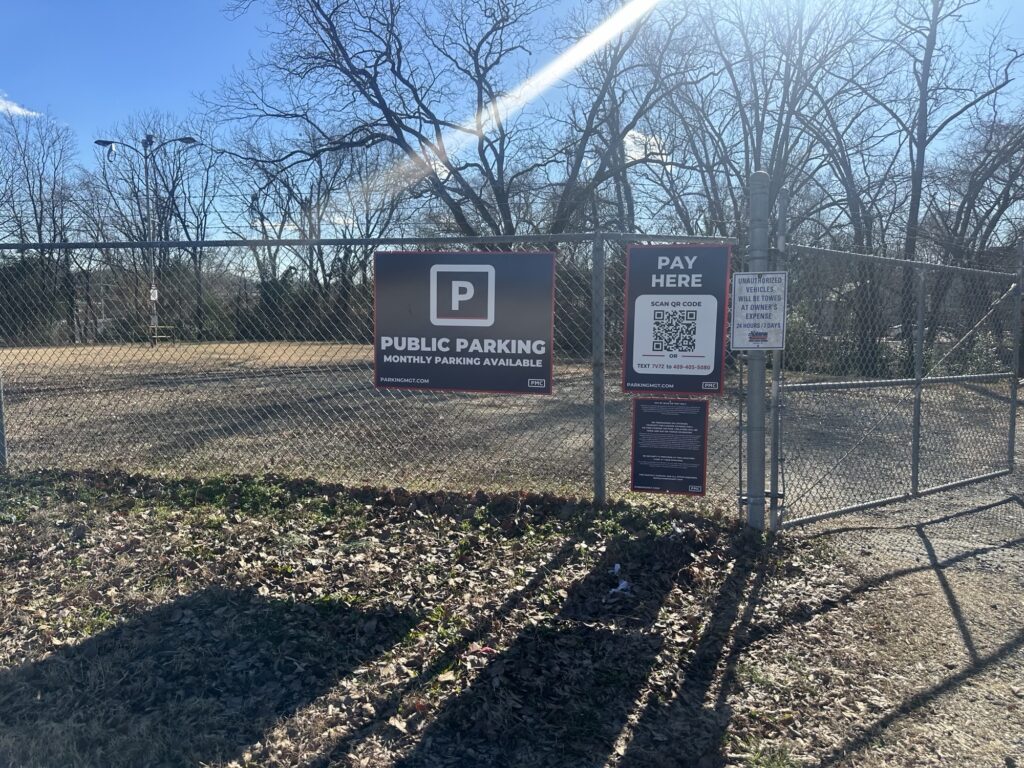 A chain-link fence with signs for public parking and pay here instructions, including a QR code. Leafless trees and a clear sky are visible in the background.