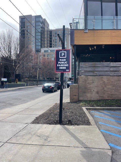 A sidewalk next to a city street with a sign reading "Paid Public Parking Here" mounted on a pole. Cars and tall buildings are visible in the background under a cloudy sky.