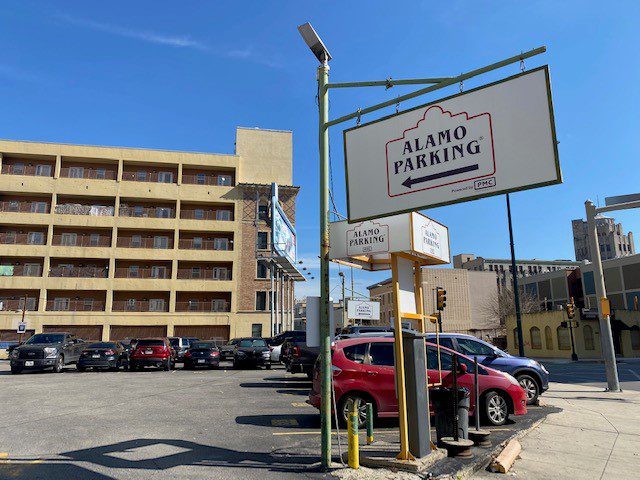 A parking lot with several parked cars is shown under a clear blue sky. A large sign reads "Alamo Parking." In the background are multi-story buildings and more parked cars.