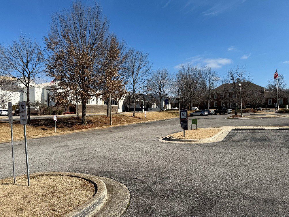 A nearly empty parking lot bordered by leafless trees and brown grass, with a few buildings and a visible American flag in the background under a blue sky.