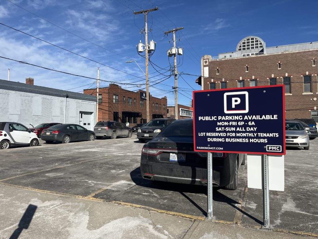 A parking lot with several parked cars on a sunny day. A red and black sign in the foreground displays public parking information and restrictions, with brick and industrial buildings in the background.