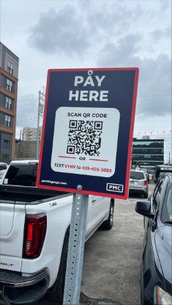 A parking lot sign reads “PAY HERE” with a QR code for payment and an option to text a code to a phone number. Parked cars and buildings are visible in the background under a cloudy sky.