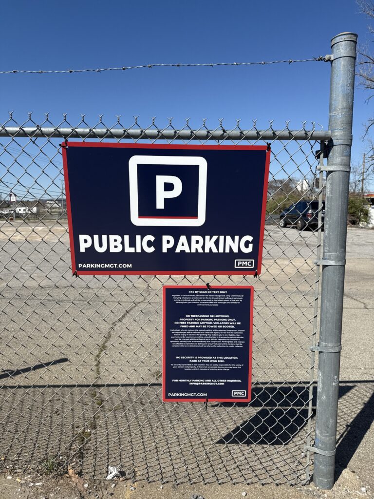 A sign on a chain-link fence reads "Public Parking" with a large "P" symbol above it. The lot behind the fence is mostly empty under a clear blue sky.
