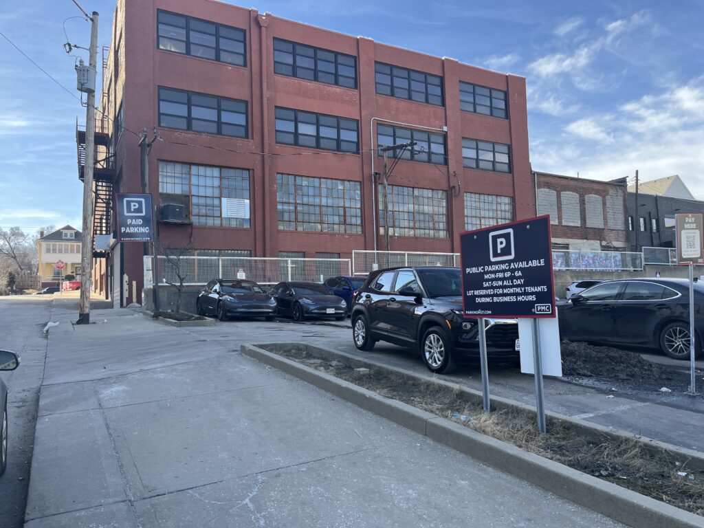 A small paid parking lot with several cars, located beside a large red brick industrial building. A sign displays parking information, and the sky is clear and blue.