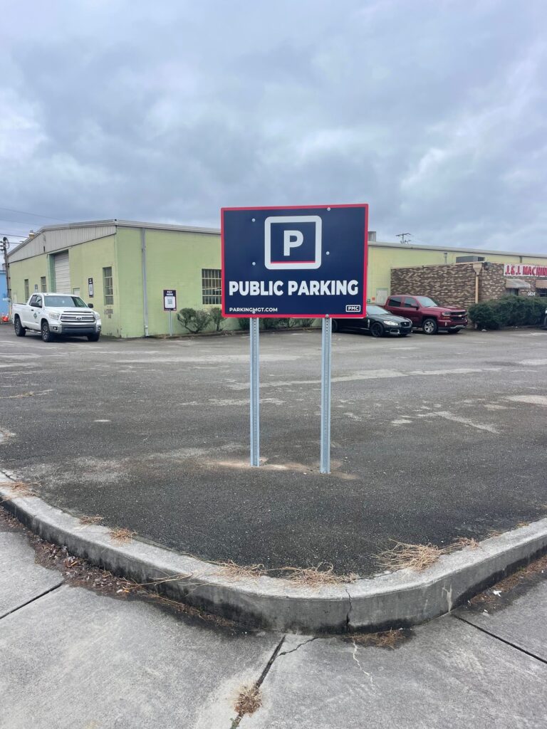 A public parking lot with a sign reading "Public Parking" on two poles, set on an empty paved area near a green industrial building and several parked vehicles under a cloudy sky.