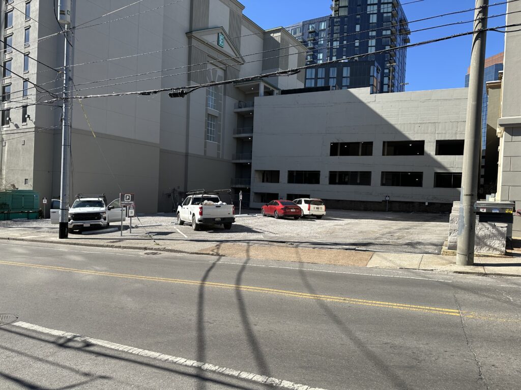 A mostly empty gravel parking lot with three parked vehicles is next to a large, light gray building. Power lines run above the street in the foreground, and tall buildings are visible in the background under a clear blue sky.