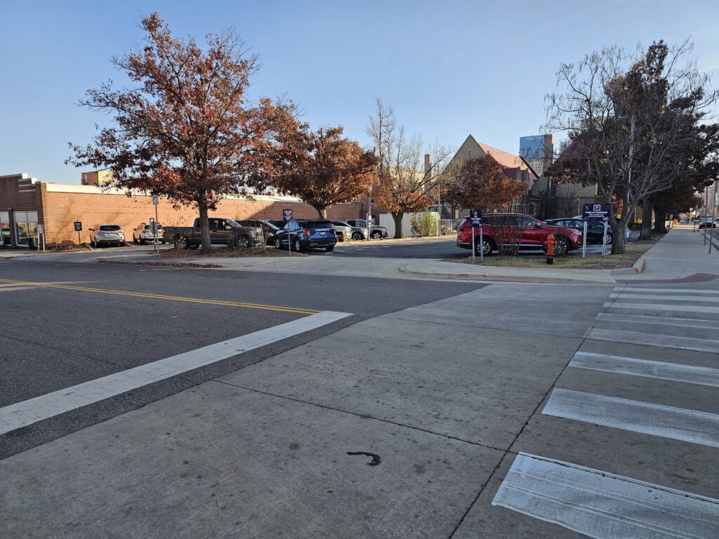 A city street with a crosswalk in the foreground, parked cars along the road, and trees with reddish-brown leaves. Buildings are visible in the background under a clear sky.