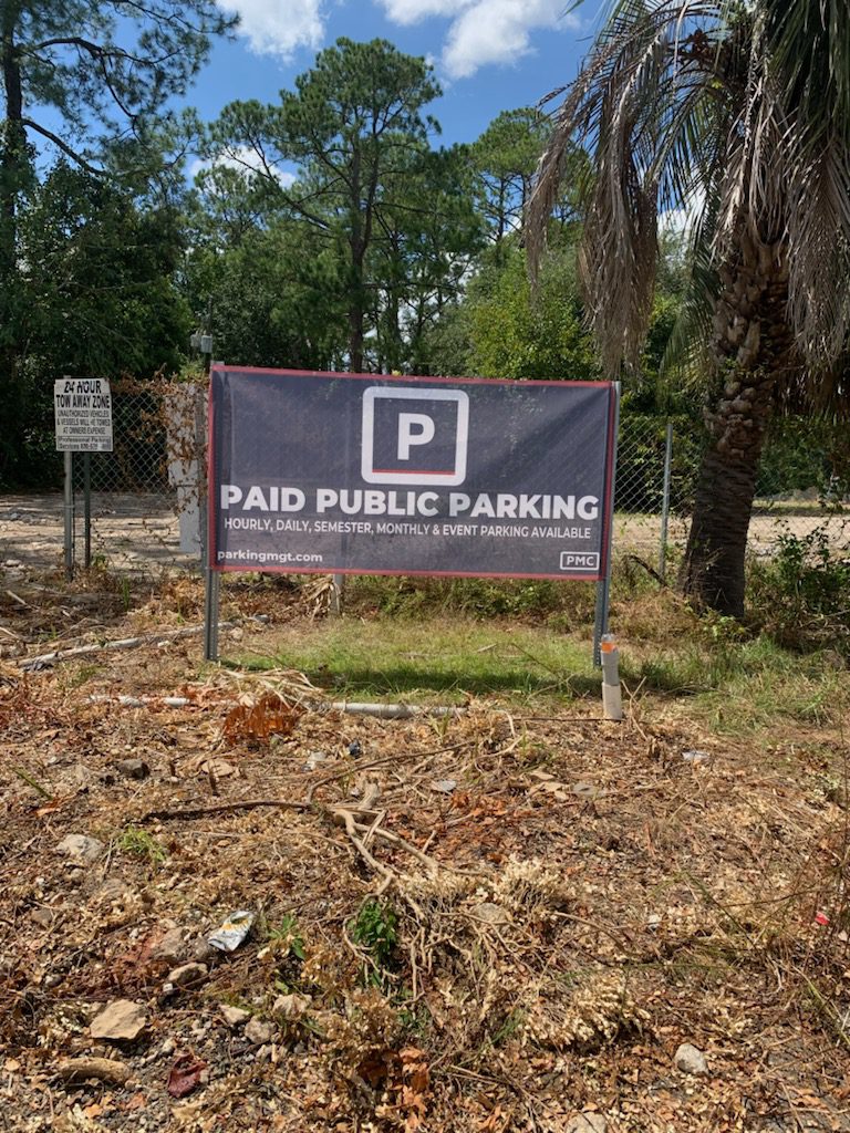 A Parking Management Company sign in a grassy lot reads "Paid Public Parking—Hourly, Daily, Semester, Monthly & Event Parking Available" with trees in the background.