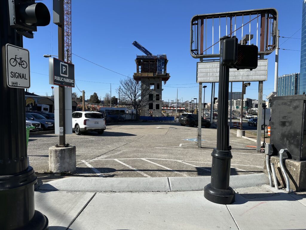 A city street scene shows a construction site with a building in progress, surrounded by parked cars and parking signs. A bike signal button and traffic lights are visible in the foreground under a clear blue sky.