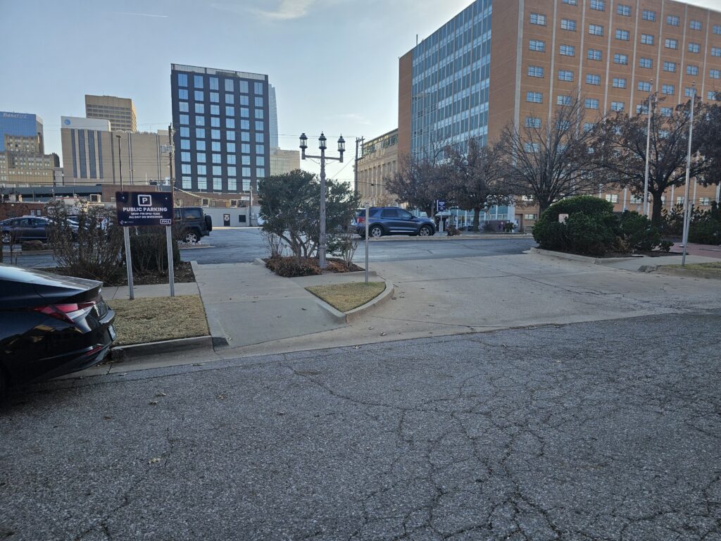 A city parking lot with a "Private Parking" sign, several cars parked, and tall office buildings in the background. Trees and street lamps line the lot, and the sky is clear.