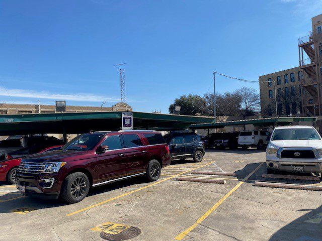 A PMC-owned parking lot with several vehicles, including a maroon SUV, a black SUV, and a white truck. Covered parking spaces, yellow lines, a sign, and a sunny sky are visible in the background.