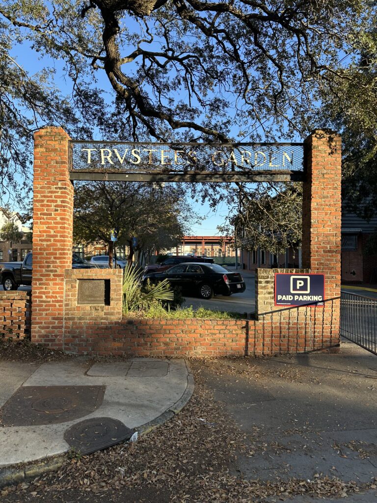 A brick entrance with a metal sign reading "Trustee Garden." A parked car and trees are visible inside. A PMC "Paid Parking" sign is attached to the gate.