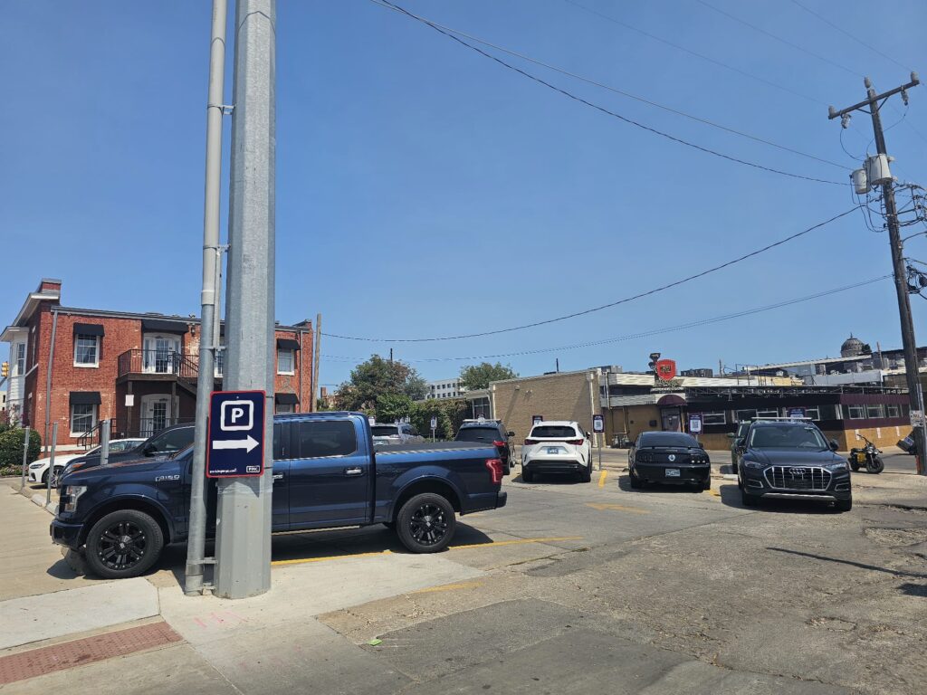 A small parking lot with several cars parked, including a black truck near a pole with a parking sign. Brick and industrial buildings are visible in the background under a clear blue sky.