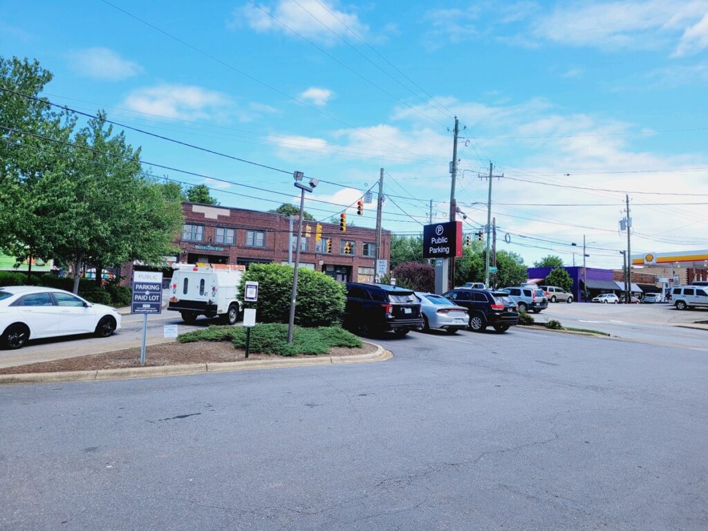 A public parking lot with several parked cars on a sunny day. A brick building and trees are in the background, with traffic lights and power lines above. Signs indicate public parking and parking payment.