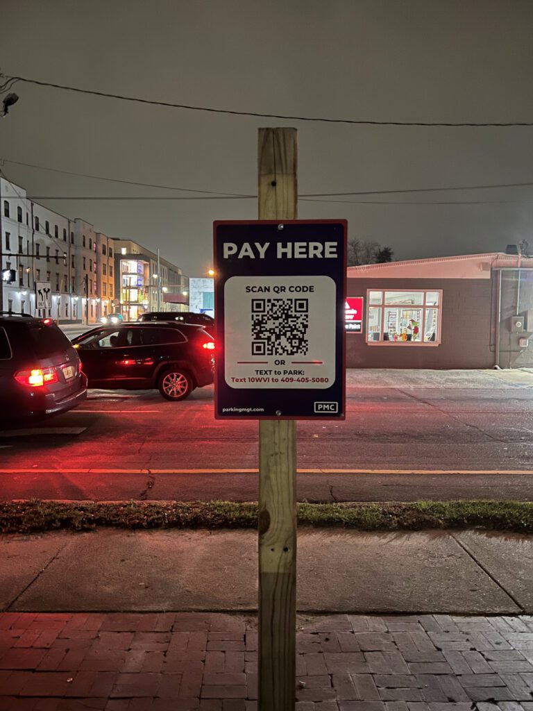 A PMC parking payment sign on a wooden post reads "Pay Here" with a QR code and parking instructions. Cars are parked nearby and buildings, including a lit store, are visible across the street at night.
