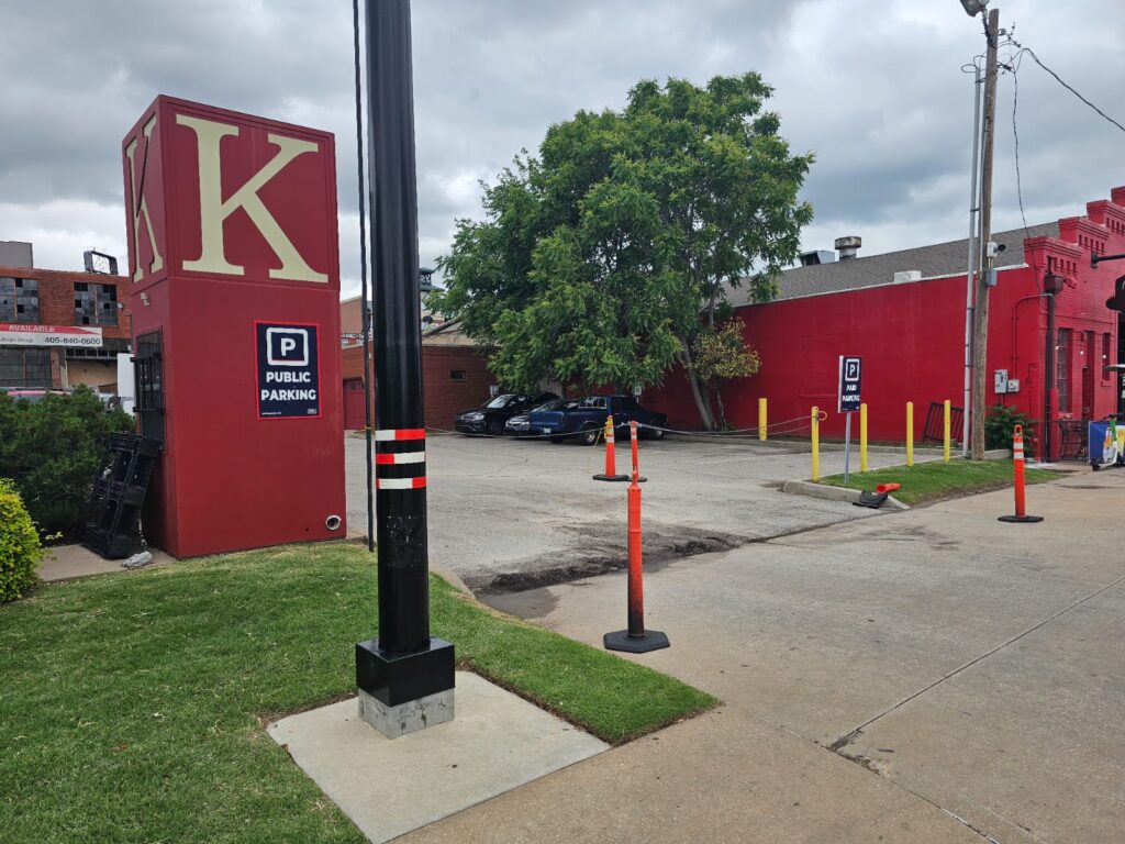 A small public parking lot bordered by red buildings, a large “K” sign, orange traffic cones, and a leafy green tree on a cloudy day. Several parked cars are visible in the background.