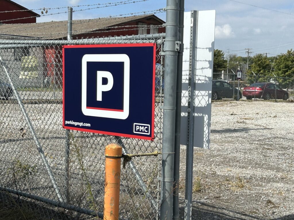 A chain-link fence with a sign showing a white "P" on a blue background, indicating parking. Behind the fence is a gravel parking lot with a few cars and some trees in the distance.
