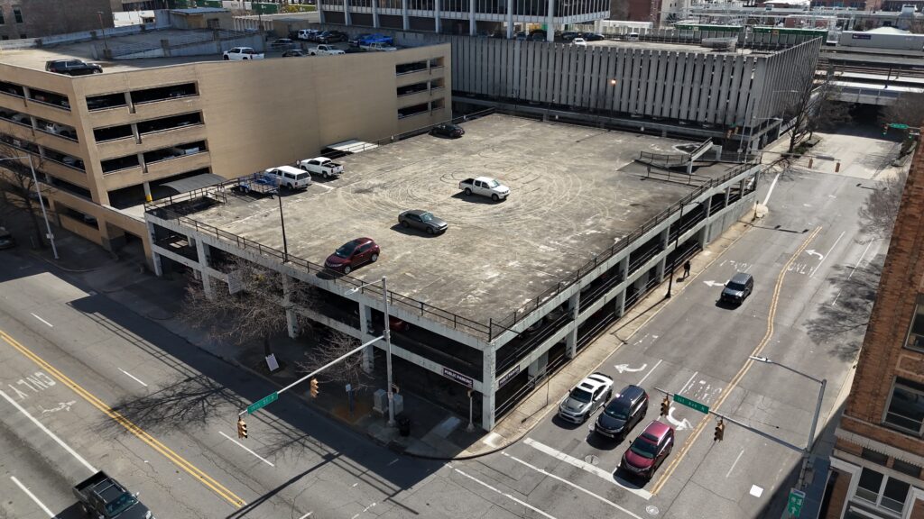 A multi-level parking garage with several cars parked on the rooftop, located at a street intersection in a city. Some trees and additional buildings surround the garage. The streets appear mostly empty.