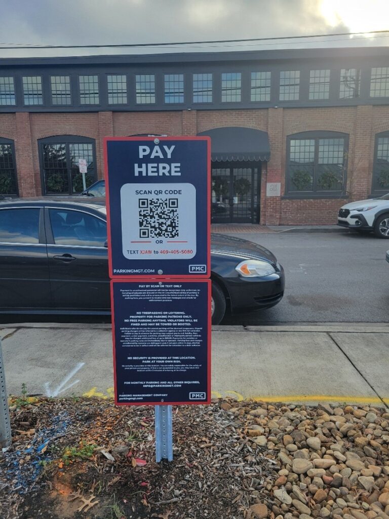 A parking payment sign on a sidewalk reads "Pay Here" and displays a QR code and payment instructions. Cars are parked on the street, and a brick building with large windows is in the background.