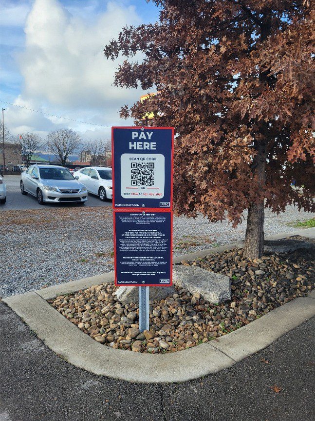 A parking lot pay station sign reads “Pay Here” with a QR code for payment, instructions beneath, and cars parked on gravel in the background next to a large tree.